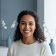 A smiling person in their late 20s to early 30s looks directly at the camera, with a soft, blurred background in blue and whi